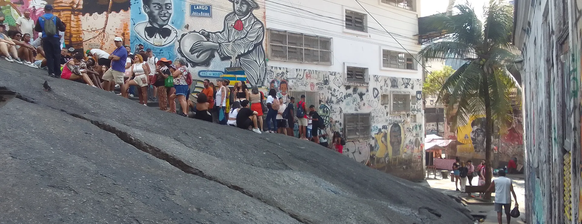 People gathered on the sloping granite rock and steps at Pedra do Sal beside a wall of murals and a palm tree