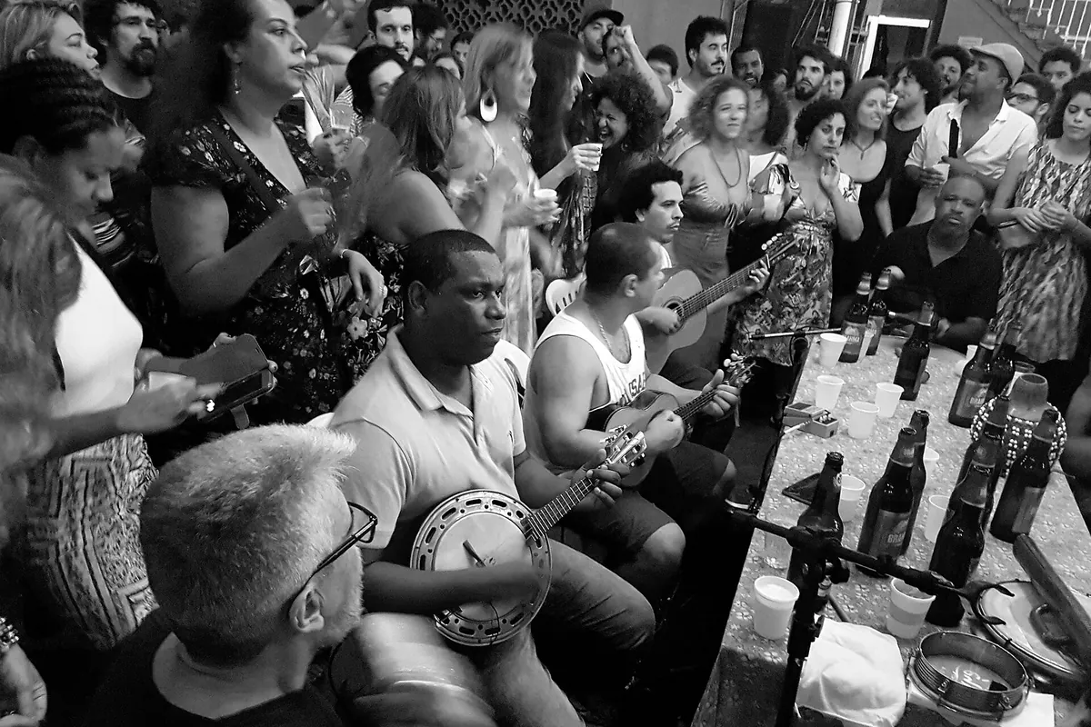Black and white photograph of musicians around a table with a banjo and a tamborim and a crowd singing, at a roda de samba in Tijuca