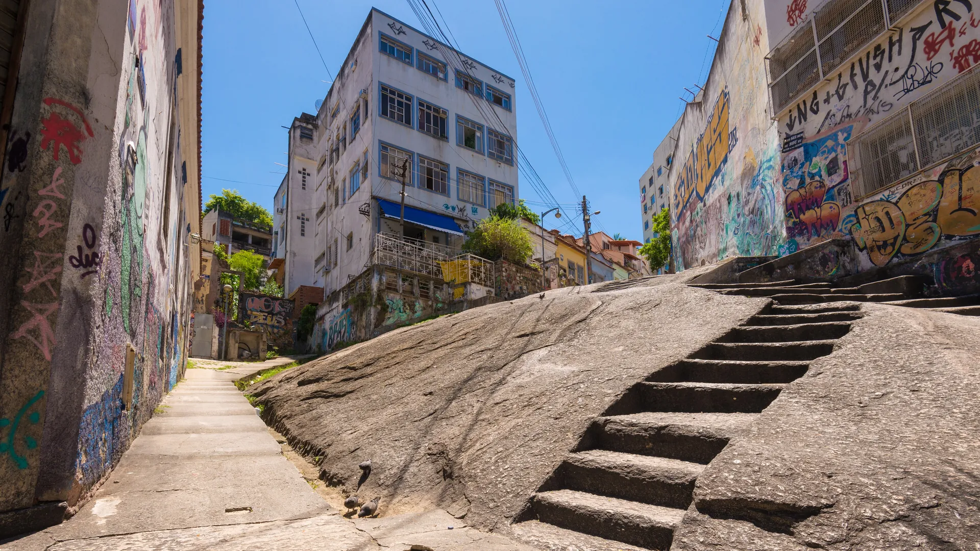 The carved granite steps of Pedra do Sal climbing a sloping rock between painted walls and apartment buildings under a clear blue sky